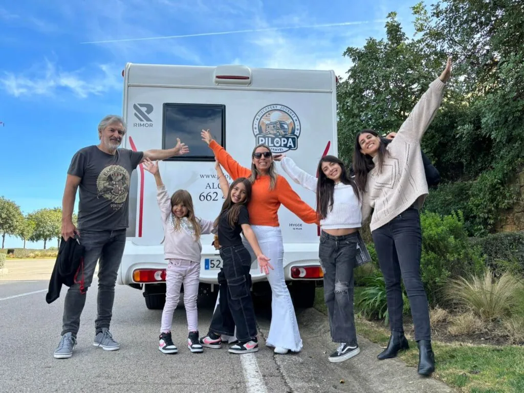 Una familia feliz de seis personas posando con los brazos abiertos frente a su autocaravana de alquiler de la marca Rimor durante un viaje.
